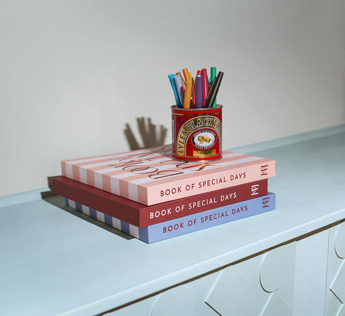 Three Book of Special Days journals stacked on a sideboard with a tin of pens